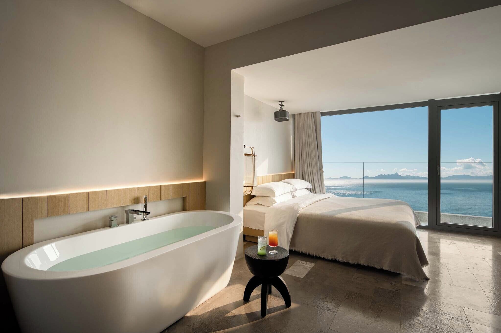 Ein modernes Hotelzimmer mit einer großzügigen Badewanne, einem Bett mit Meerblick und einem kleinen Tisch mit einem Getränk.