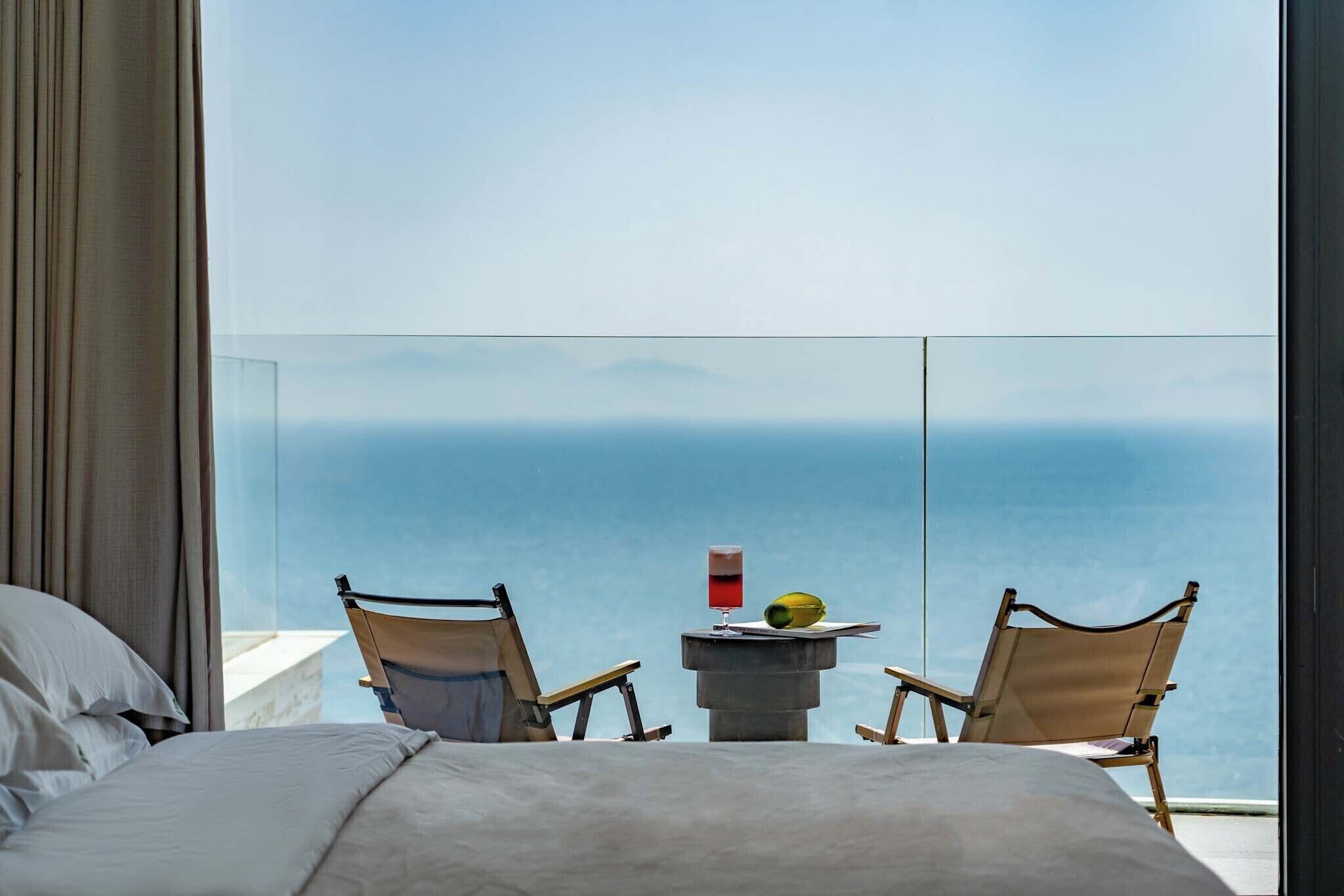A bedroom with a large bed, two chairs, a small table with a drink and fruit, and a view of the ocean.
