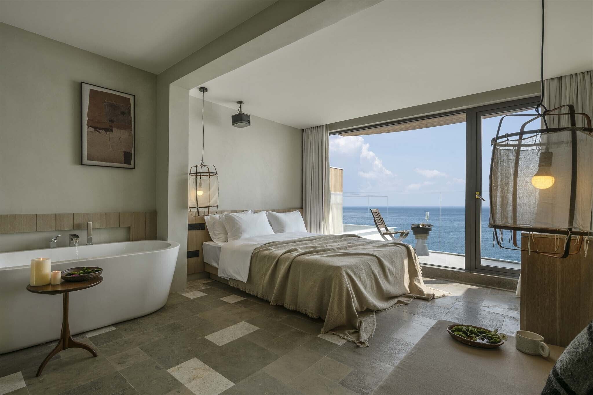 A modern bathroom with a freestanding bathtub, a bedside table with candles, and a view of the sea through large windows.