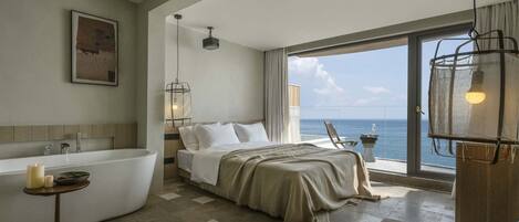 A modern bathroom with a freestanding bathtub, a bedside table with candles, and a view of the sea through large windows.
