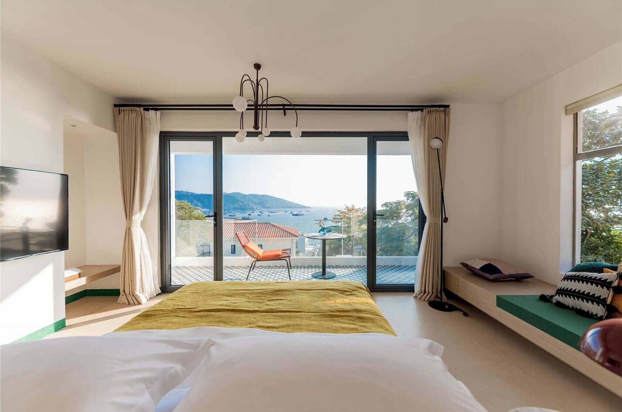 Une chambre avec un lit, une télévision, une fenêtre avec des rideaux, une vue sur l’extérieur et un balcon avec une chaise et une table.