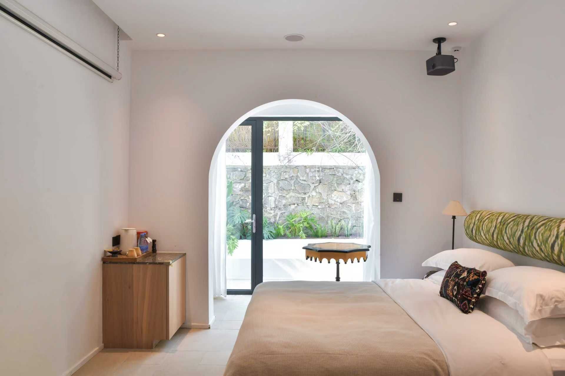 A bedroom with a bed, a bedside table, a lamp, and a view of a garden through an arched doorway.