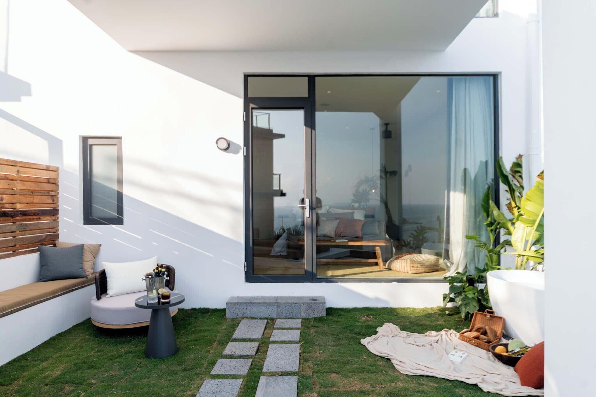 Un área de estar moderna al aire libre, con un banco acolchado, una mesa redonda y un ventanal de vidrio que da a una habitación.