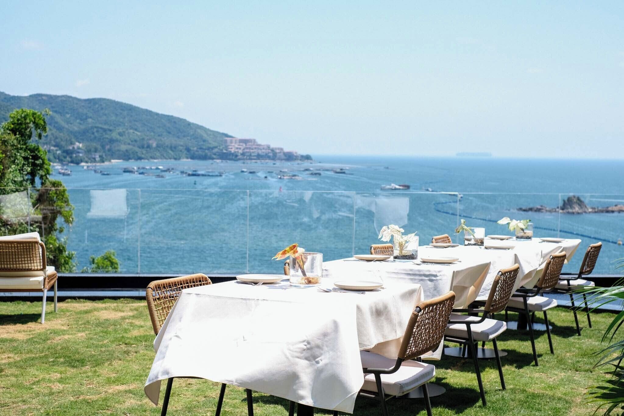 Un espacio de comedor al aire libre con mesas dispuestas para una comida, con vista a la costa, barcos y colinas.