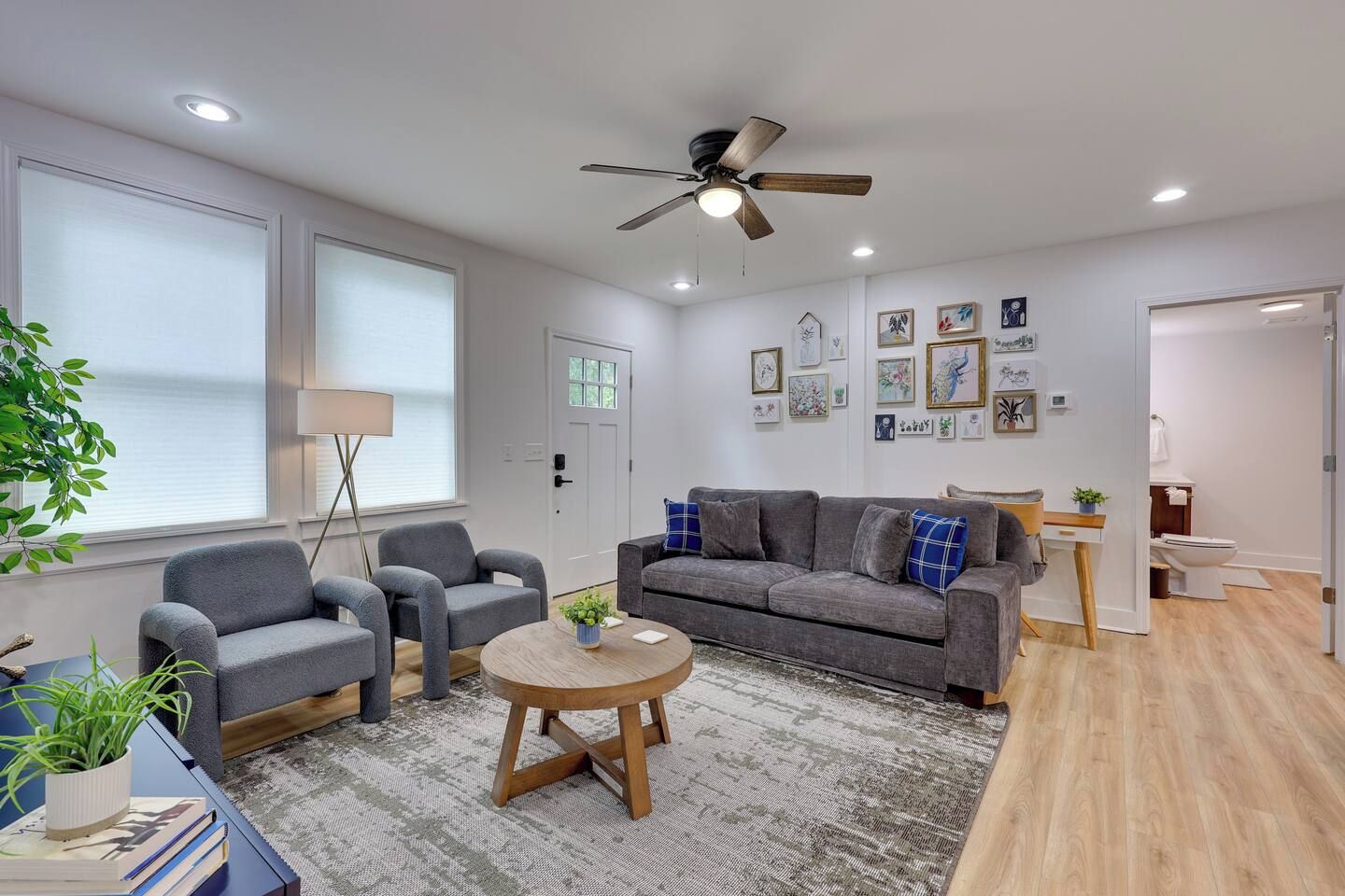 A modern living room with a grey sofa, two armchairs, a coffee table, and a ceiling fan. There are framed pictures on the wall and a door leading to a bathroom.