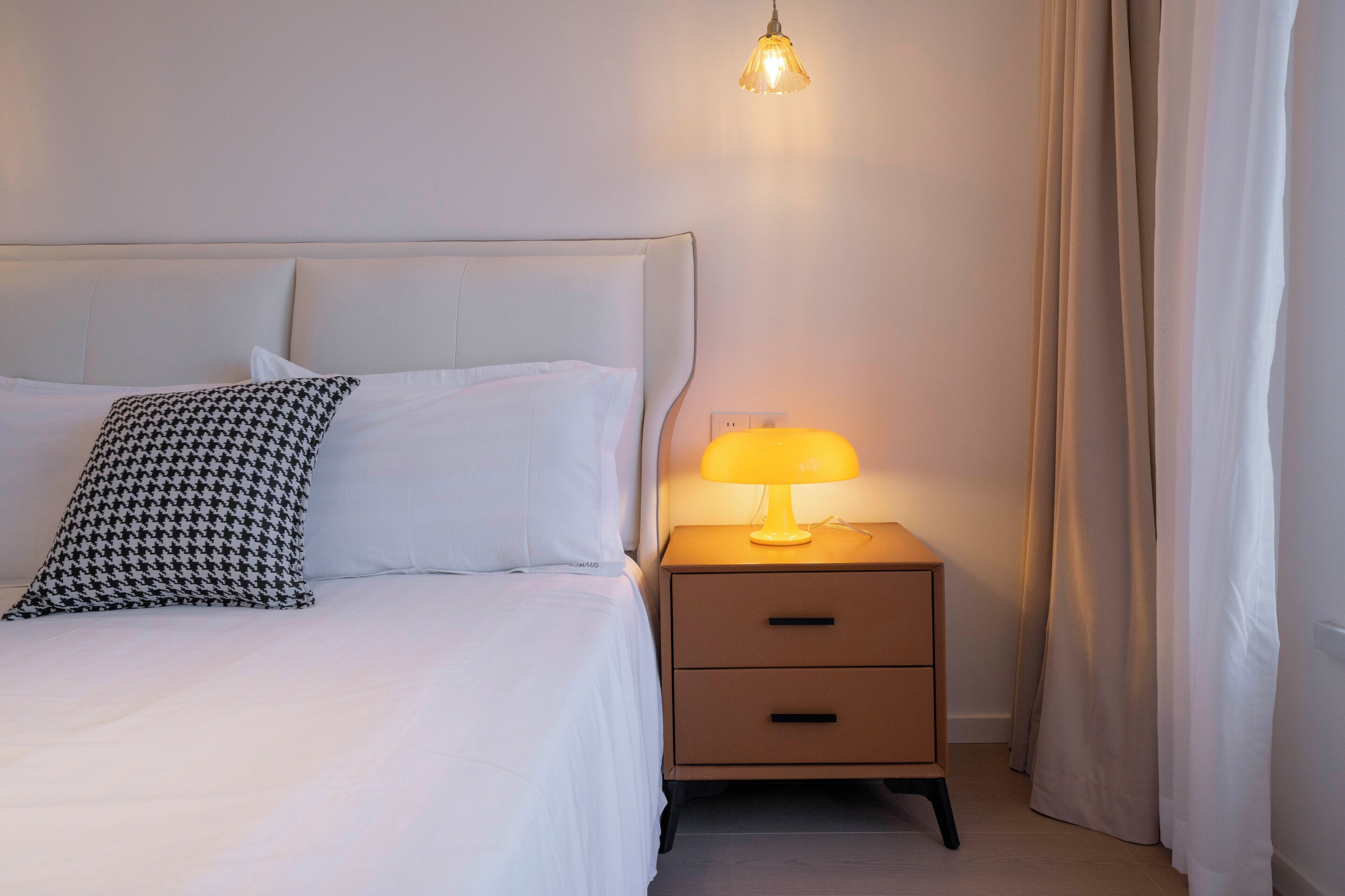 Ein ordentlich bezogenes Bett mit weißen Leinen und einem Hundszahn-Kissen, ein Nachttisch mit Lampe sowie eine hängende Leuchte.