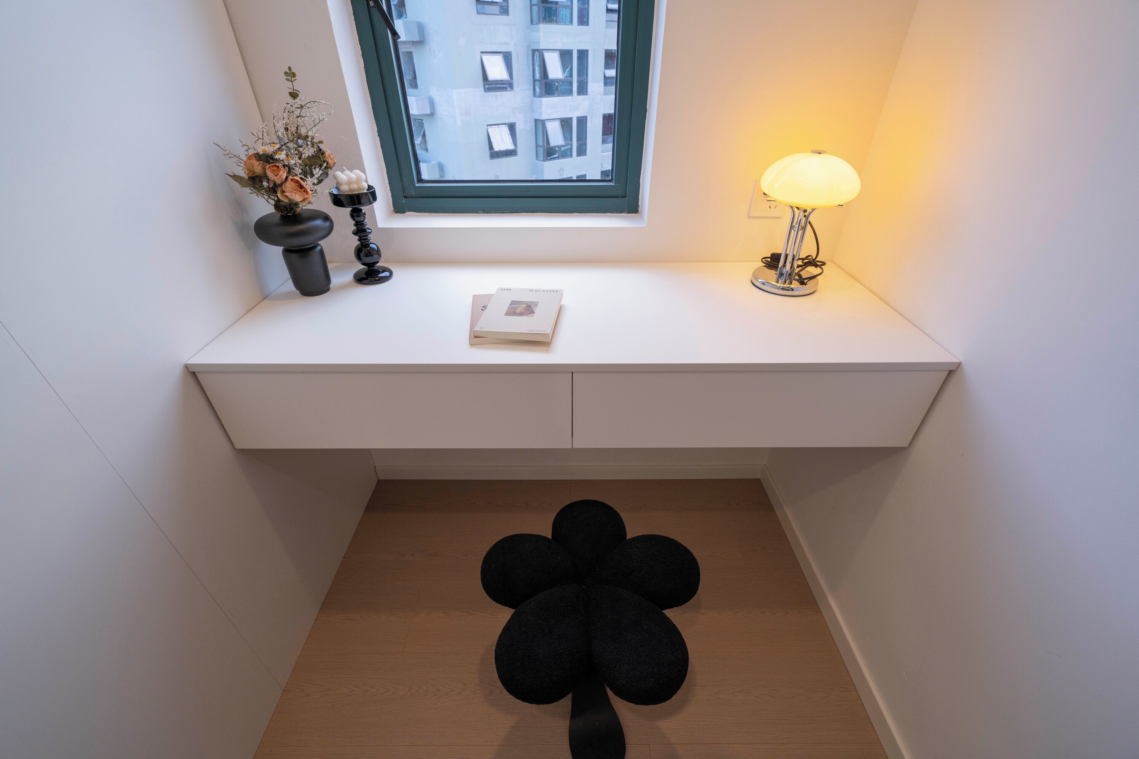 A small, white desk with a window, a lamp, a vase with flowers, and a black decorative item on the floor.