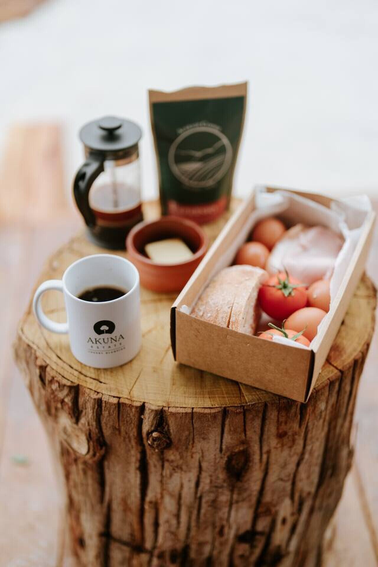 A rustic wooden table with a coffee pot, a mug labeled "AKUNA," a small bowl, and a cardboard box containing eggs and tomatoes.