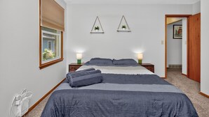 Ein Schlafzimmer mit einem großen Bett, Nachttischlampen, einem Fenster mit Jalousien und zwei Wandregalen mit Pflanzen.