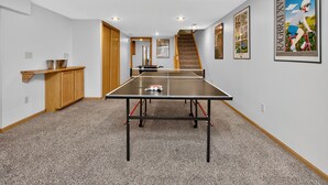 Ein Tischtennis-Tisch in einem Raum mit Treppe, Holzschränken und gerahmten Bildern an den Wänden.