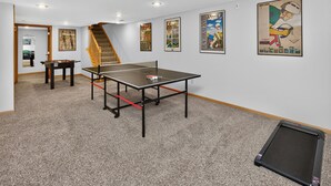 Ein Tischtennis-Tisch in einem Raum mit Teppichboden, einer Treppe und mehreren gerahmten Bildern an den Wänden.