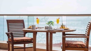 Une table ronde en bois, accompagnée de deux chaises, d’une petite plante en pot et de deux verres de jus d’orange décorés de cerises, le tout placé sur un balcon offrant une vue sur la mer.