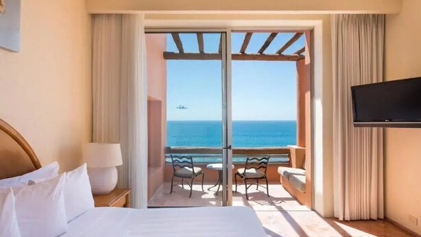 Une chambre d’hôtel avec un lit, une télévision, un balcon avec des chaises et une vue sur la mer.