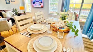 A dining area with a wooden table set for four, a television mounted on the wall, and a view of the outdoors through large windows. - Wonderful apartment 10 minutes from Disney (Kissimmee)
