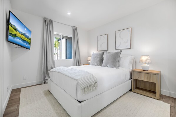 A bedroom with a bed, a TV mounted on the wall, a bedside table with a lamp, and two framed pictures on the wall. - Rancho Haven (Tarzana)