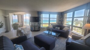 A spacious living room with a large window overlooking the beach, furnished with dark sofas, a coffee table, and a flat-screen TV.