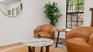 A room with two leather armchairs, a round marble table with a potted plant, a mirror on the wall, and a window with a view of trees.