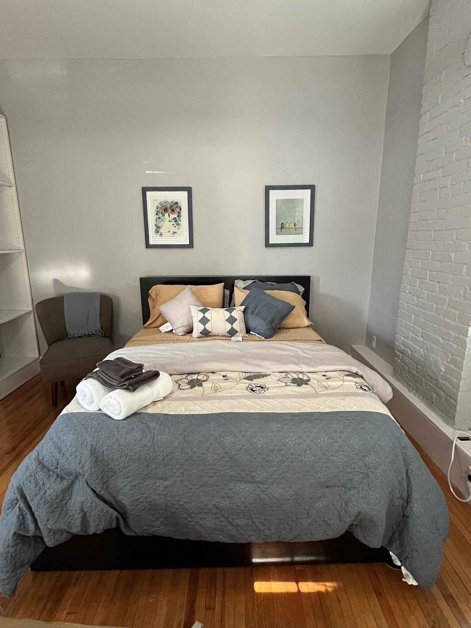 A neatly made bed with a grey and white duvet, a chair, and two framed pictures on the wall.