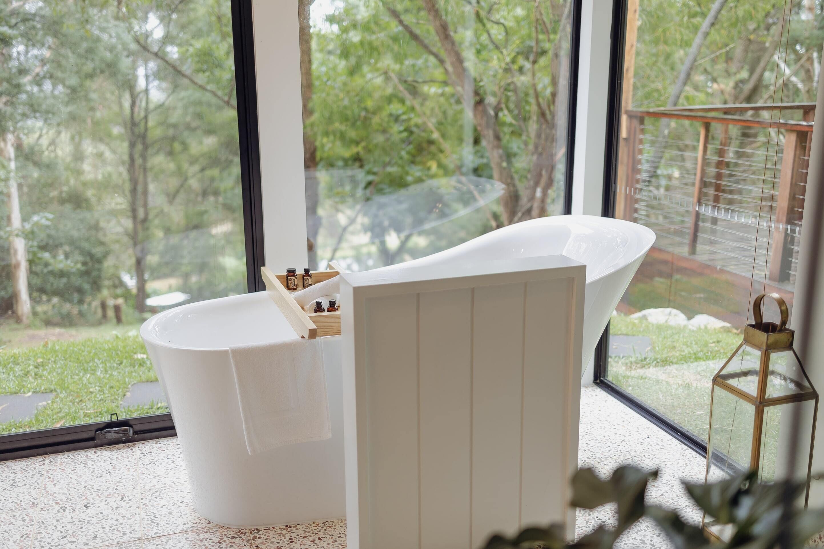 Een moderne badkamer met een vrijstaand bad, een houten nachtkastje met toiletartikelen en uitzicht op een tuin door grote ramen.