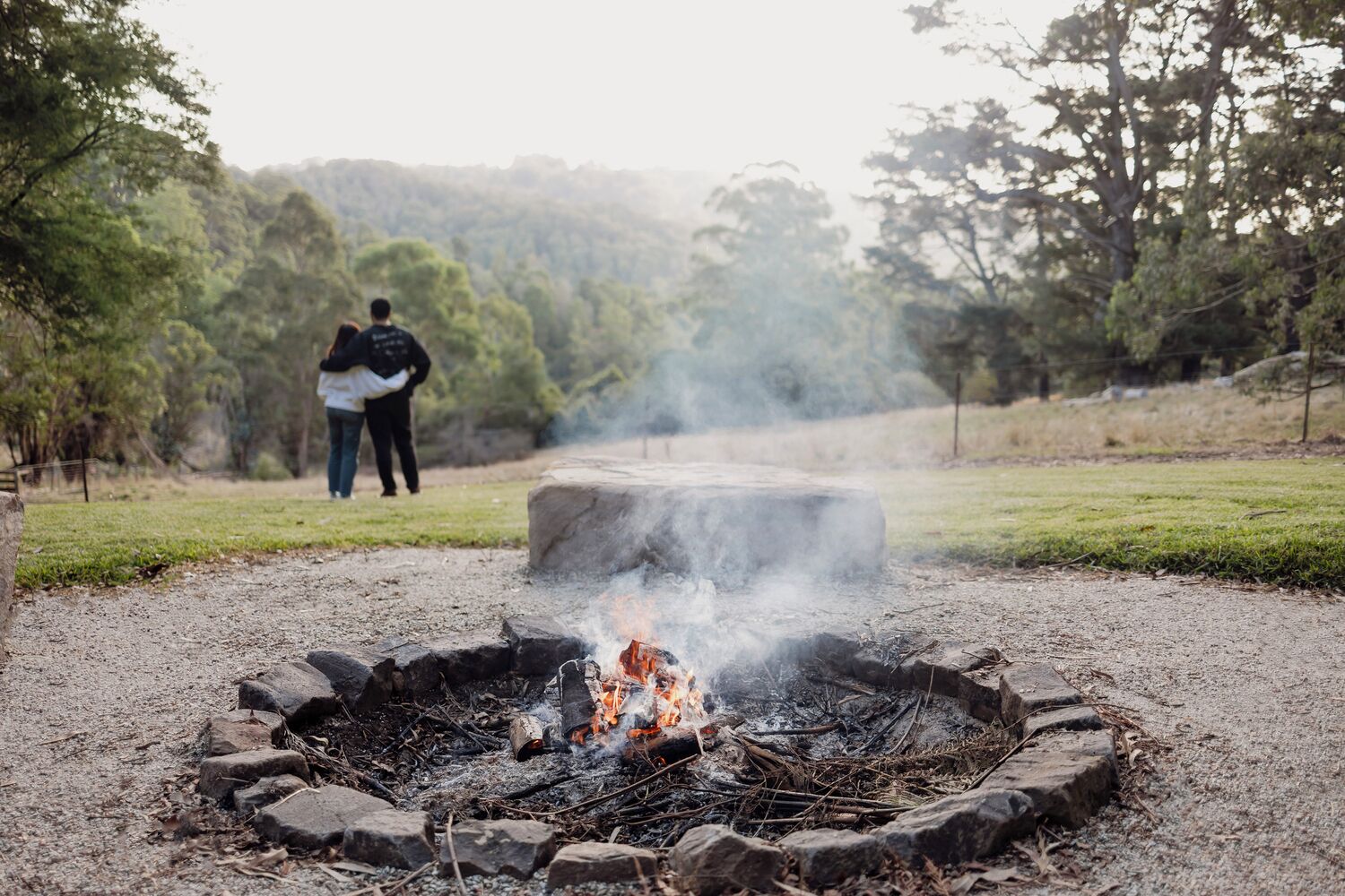 A campfire with rocks arranged in a circle, surrounded by a grassy area with trees in the background.