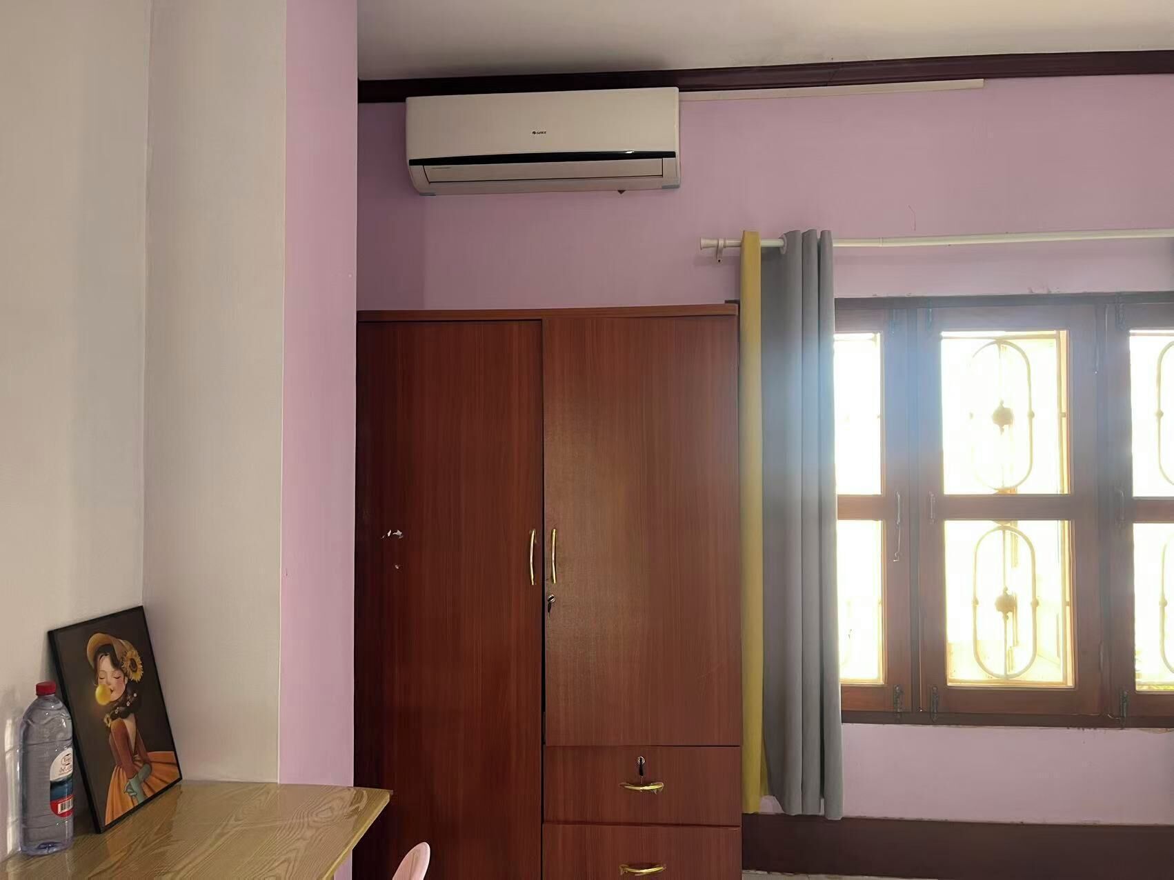 A room with a wooden wardrobe, an air conditioning unit, a table with a picture frame and a bottle, and a window with curtains.
