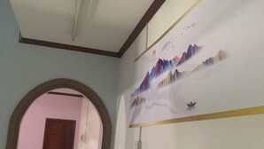 Un couloir se terminant par une porte en bois, un mur blanc orné d’un tableau représentant un paysage et un sol carrelé.