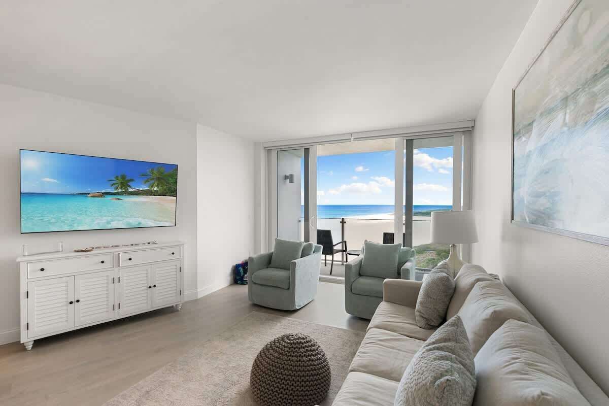 A modern living room with a large flat-screen TV, a white console, a beige sofa, and a view of the beach through large windows.