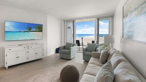 Een moderne woonkamer met een grote flatscreen televisie, een witte console, een beige bank en uitzicht op het strand door grote ramen.