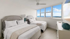 Een slaapkamer met twee bedden, een plafondventilator, een nachtkastje, een dressoir en een groot raam met uitzicht op de lucht en wolken.