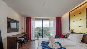Hotellihuone, jossa on sänky, työpöytä, televisio ja parveke, jossa on verhot.