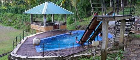 A swimming pool with a slide, surrounded by a wooden deck and a covered pavilion.