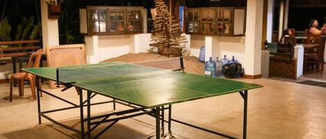 Una mesa de ping pong en una habitación con suelo de hormigón, sillas de madera y un pequeño árbol de Navidad.