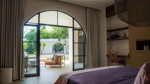 A bedroom with a large bed, a view of a garden through a glass door, and a seating area with a table and chairs. - Villa Sungai (Cepaka)