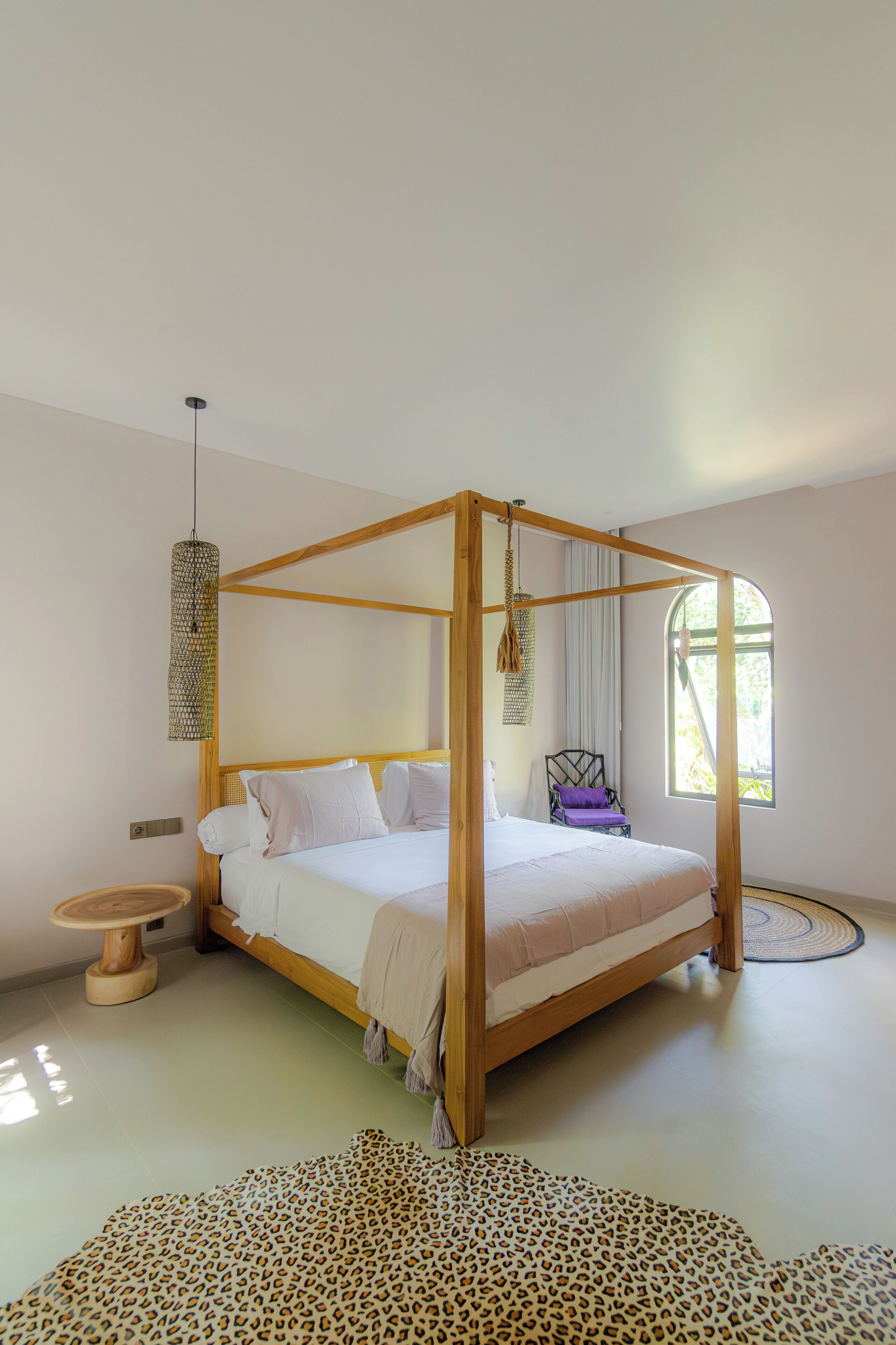 A four-poster bed with a wooden frame and white bedding, a round wooden side table, a leopard print rug, and a window with curtains.