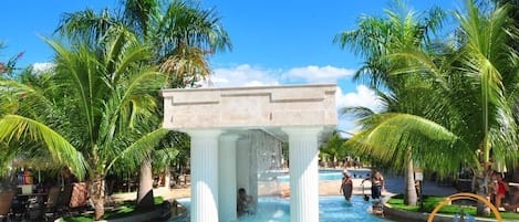 A pool area with a central fountain, surrounded by palm trees and lounge chairs.
