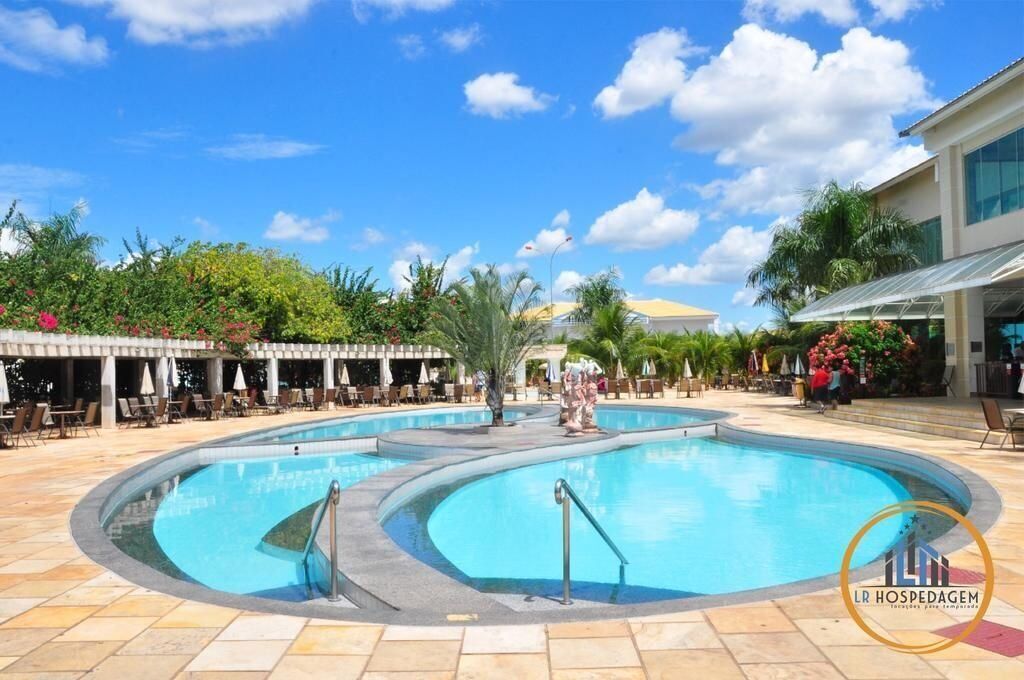 A pool area with lounge chairs and umbrellas, surrounded by greenery and a building with a glass facade.