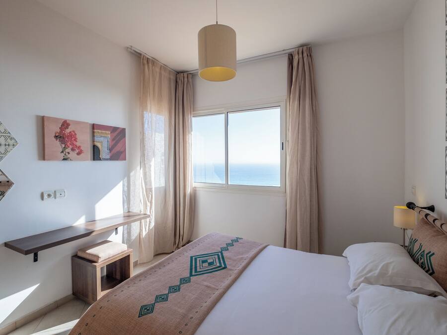 A hotel room with a bed, a bedside table, a window with curtains, a wall-mounted lamp, and a painting.