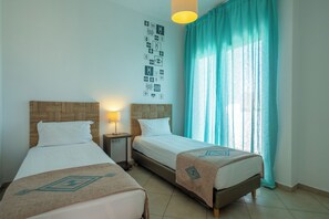 A hotel room with two beds, a nightstand, a lamp, and a large window with curtains. - Lunja Village Taghazout (Tamri)
