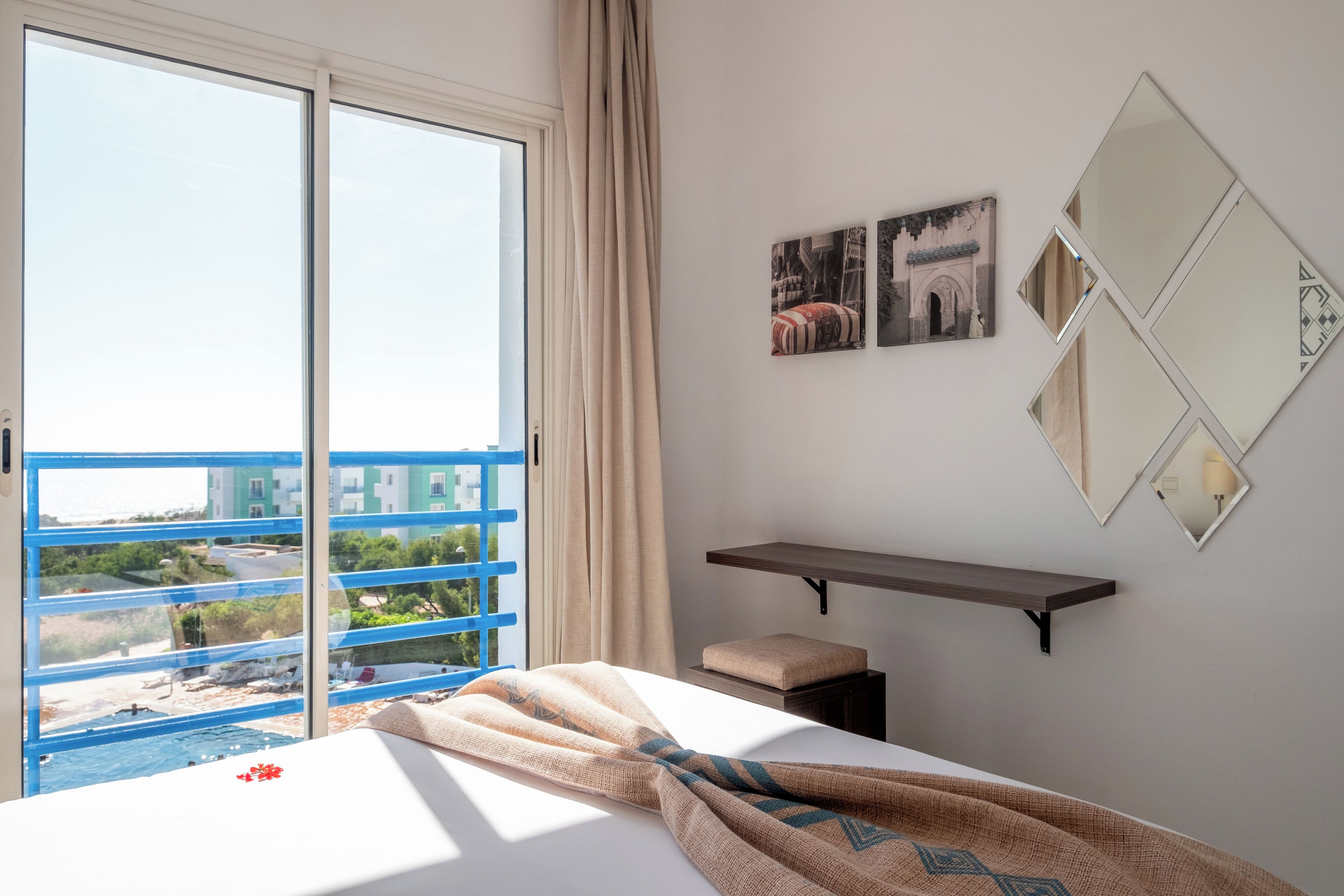 A hotel room with a large window, a bed with a blanket, a small stool, and a shelf with pictures.
