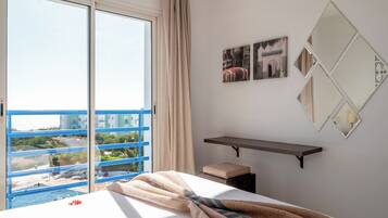 A hotel room with a large window, a bed with a blanket, a small stool, and a shelf with pictures.