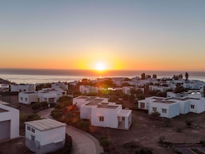 Una zona residencial costera con edificios blancos modernos, una vista despejada del atardecer sobre el mar y un camino pavimentado que atraviesa la propiedad.