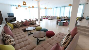 A spacious lounge area with multiple seating options, including sofas and round tables, and a television on the left.