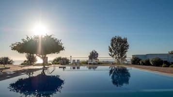 A clear sky with the sun low on the horizon, a swimming pool reflecting trees and the sky, and a fenced area with lounge chairs.