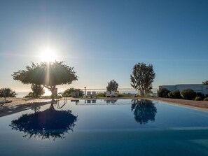 Un ciel dégagé avec un soleil bas à l’horizon, une piscine qui reflète les arbres et le ciel, et un espace clos avec des chaises longues.