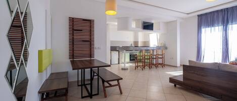 A modern kitchen with a central island, a dining area with a wooden table and benches, and a living area with a sofa and a TV.