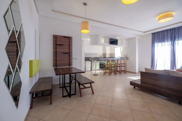 A modern kitchen with a central island, a dining area with a wooden table and benches, and a living area with a sofa and a TV. - Lunja Village Taghazout (Tamri)