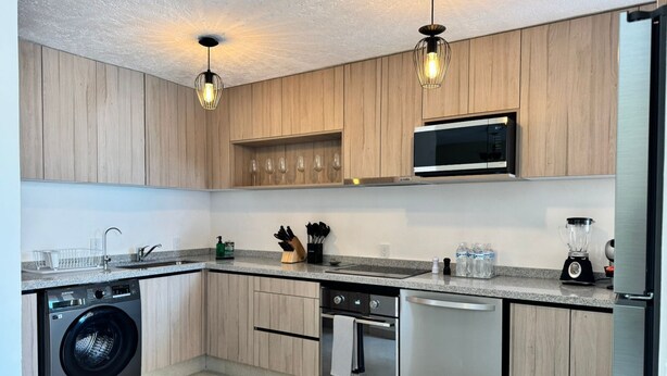 A modern kitchen with wooden cabinets, a built-in microwave, a dishwasher, a washing machine, and pendant lighting.