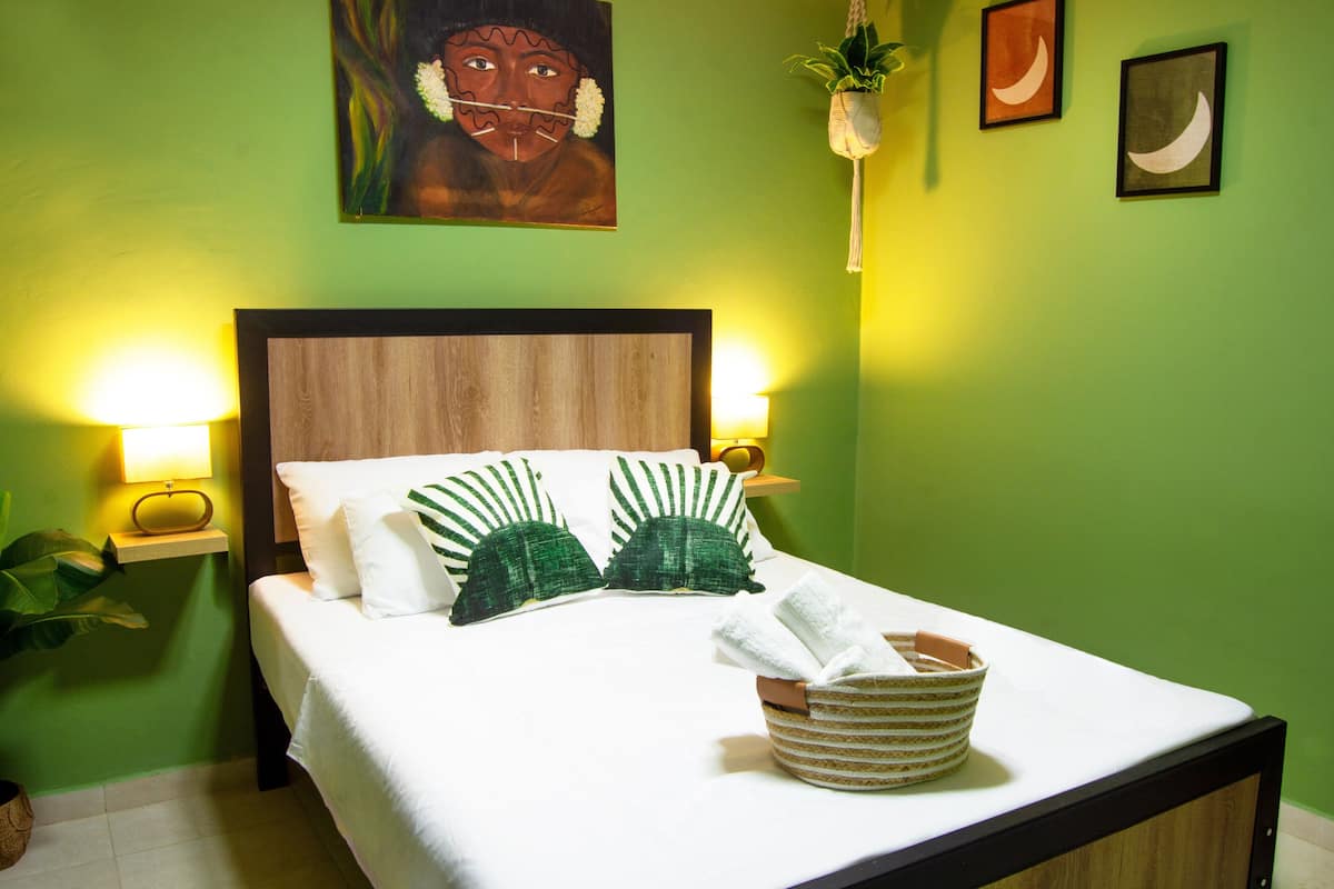 A neatly made bed with white linens and green pillows, a bedside lamp, and a woven basket on the nightstand.