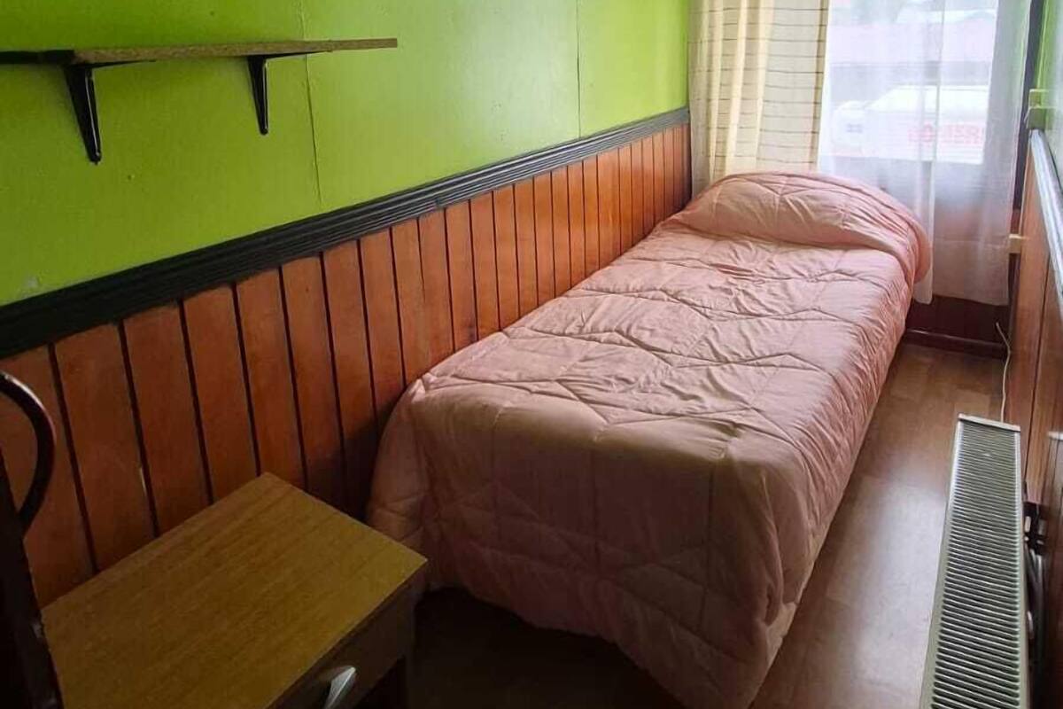 Una habitación pequeña con cama individual, un banco de madera, un radiador y una ventana con cortinas.