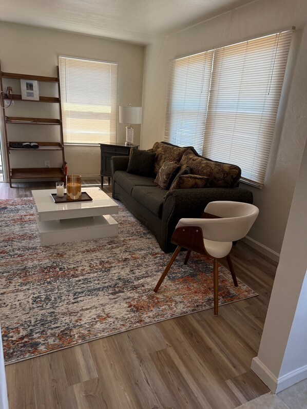 A modern living room with a dark green sofa, a white coffee table, and a patterned rug. There are two windows with blinds, a wooden shelf, and a lamp on a side table.